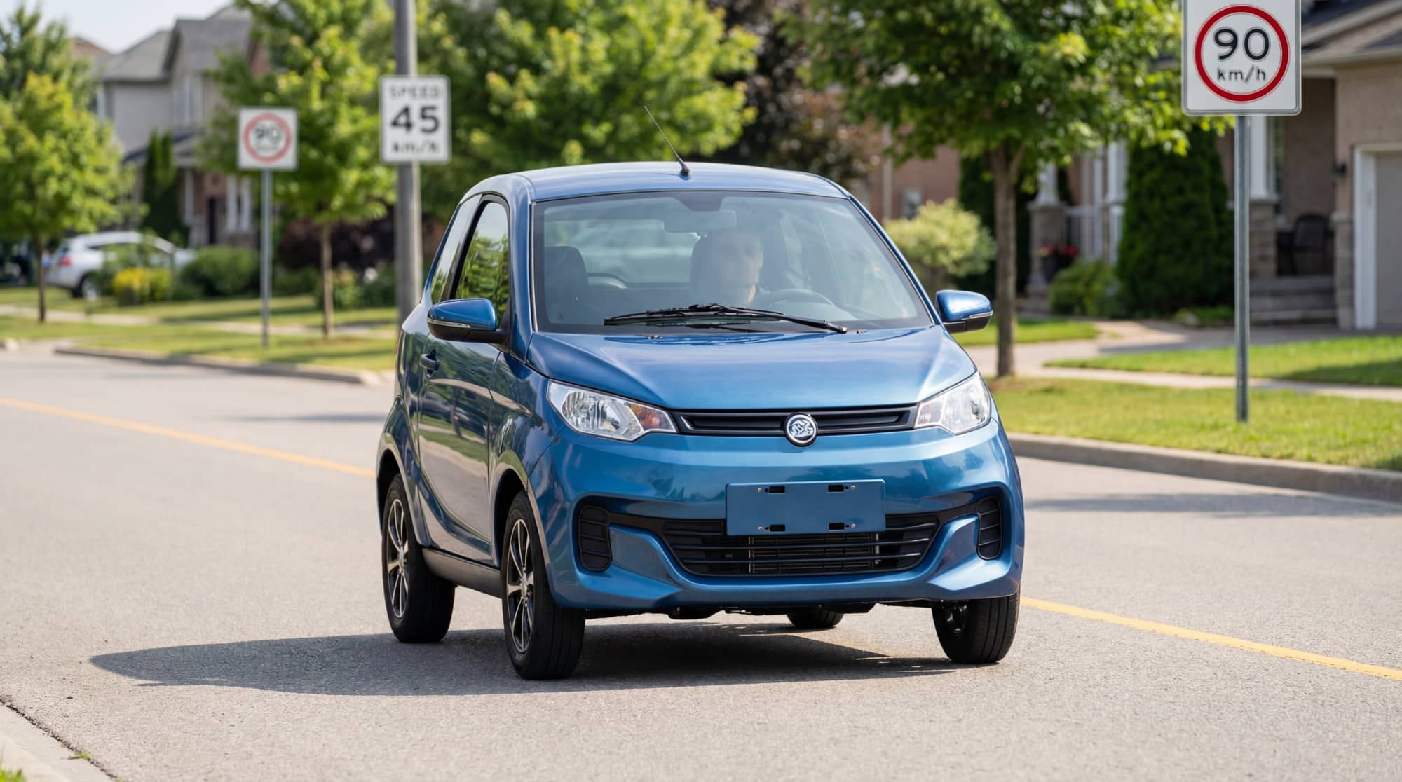 Une voiturette bleue de type sans permis roule sur une route résidentielle ensoleillée. Des panneaux indiquent 45 km/h et 90 km/h.